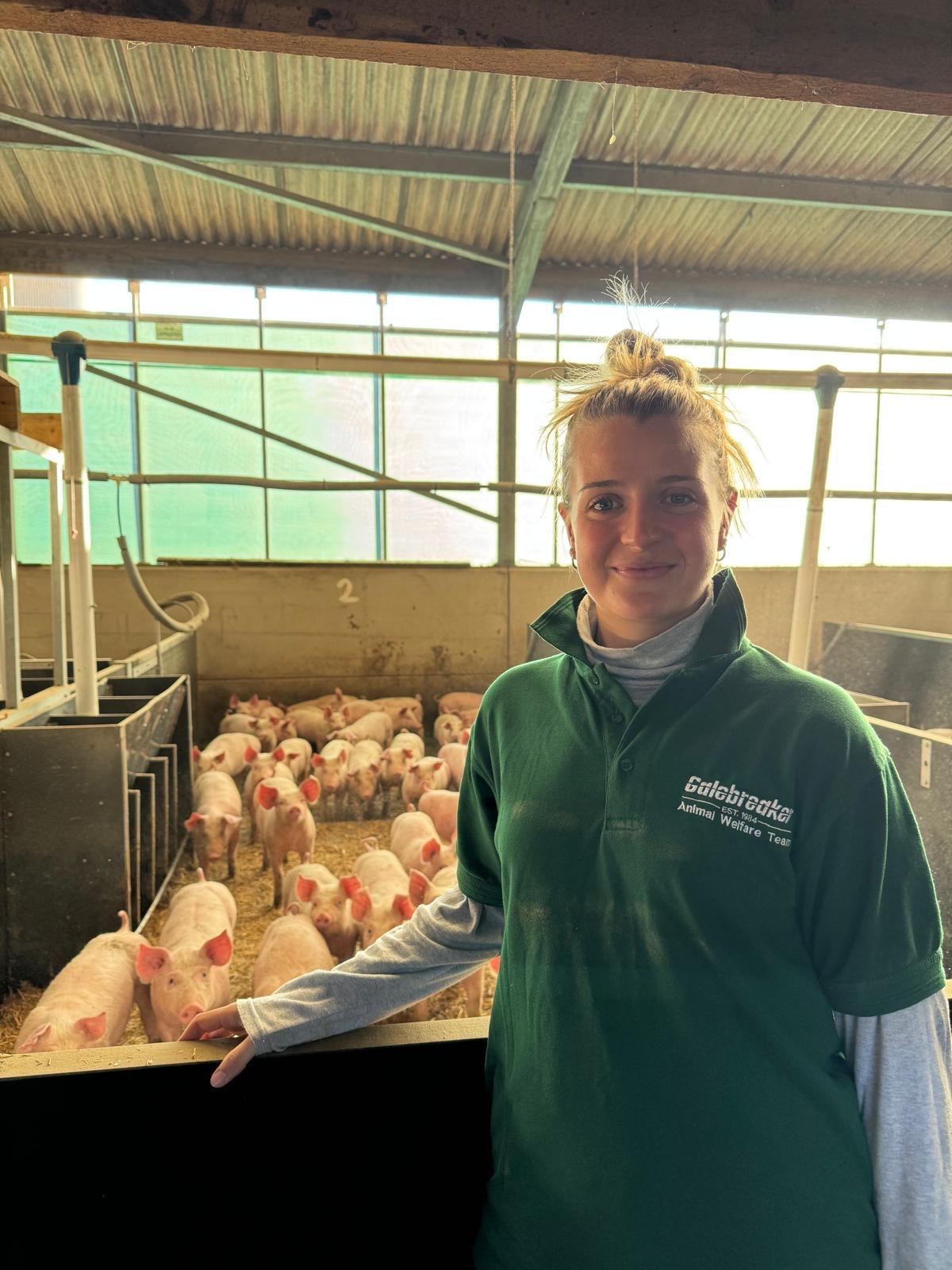 Young woman wearing green polo shirt stood in front of pen of pigs.